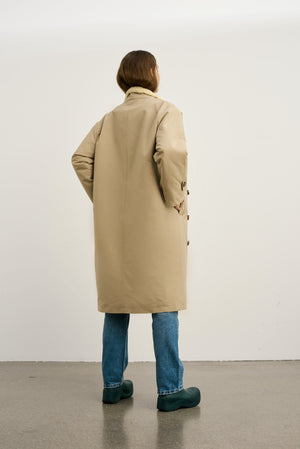 A woman stands with her back to the camera, wearing a beige MEOTINE coat, paired with blue jeans and green shoes.