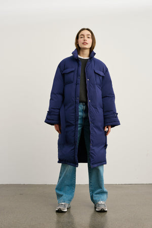 A model stands confidently in a long, navy blue puffer coat by MEOTINE, paired with relaxed jeans and sneakers.