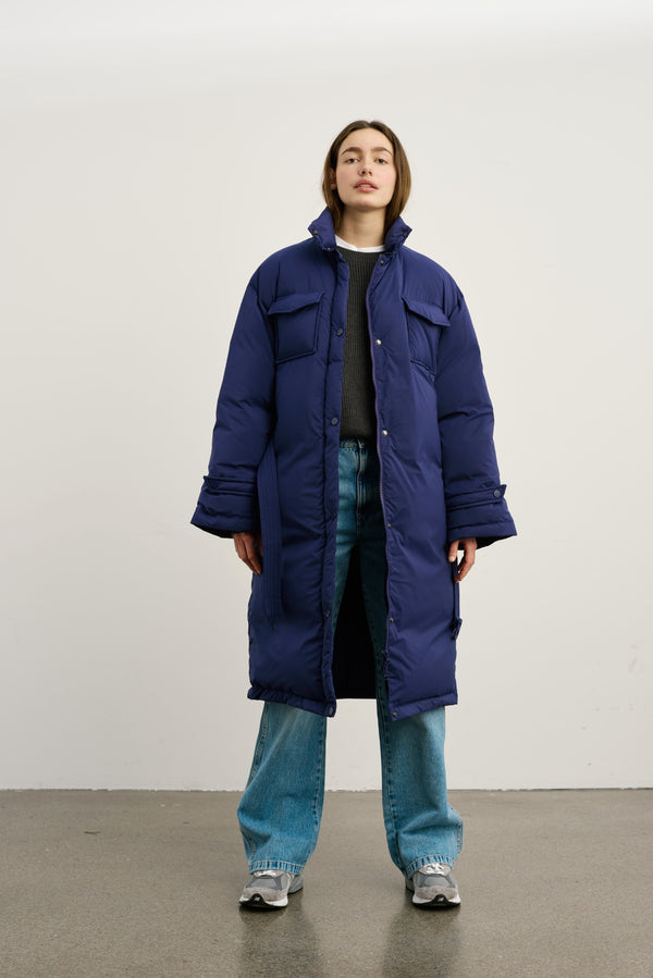 A model stands confidently in a long, navy blue puffer coat by MEOTINE, paired with relaxed jeans and sneakers.