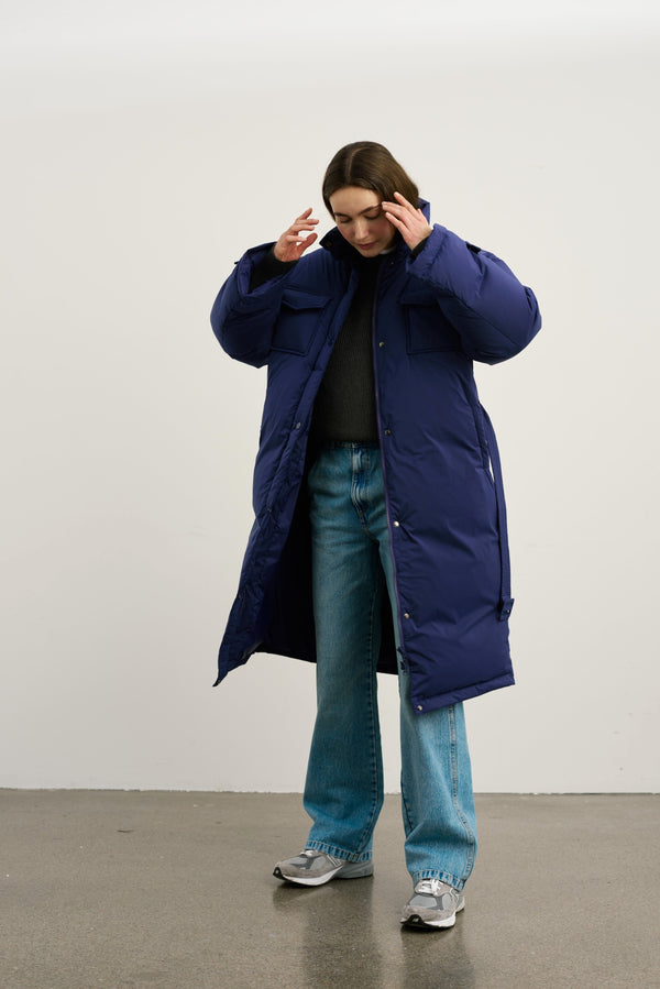A model wearing a long navy blue puffer coat from MEOTINE, paired with a dark sweater and light blue jeans, striking a thoughtful pose.