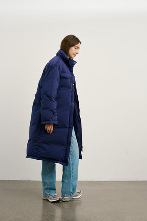 A model wearing a long navy puffer coat from MEOTINE, paired with light blue jeans and sneakers, standing against a plain background.