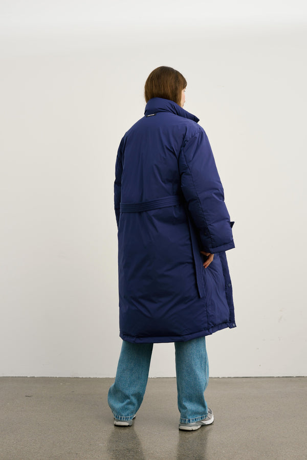 A model wearing a long navy blue MEOTINE puffer coat, standing with her back to the camera, paired with light blue jeans.