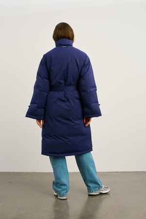 A model wearing a long navy blue puffer coat from MEOTINE, styled with light blue jeans and sneakers, standing against a plain background.
