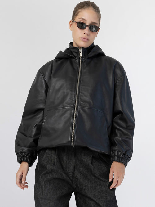 Stylish black leather jacket with a hood, featuring a zip closure and relaxed fit, paired with sleek sunglasses. MEOTINE.