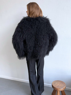 A model showcases a fluffy black MEOTINE jacket from the back, paired with sleek black trousers against a minimalist backdrop.