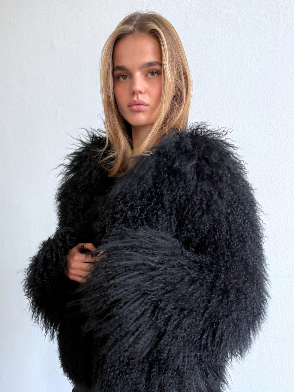 A model showcases a luxurious black fluffy coat by MEOTINE, paired with sleek black pants, set against a minimalist backdrop.