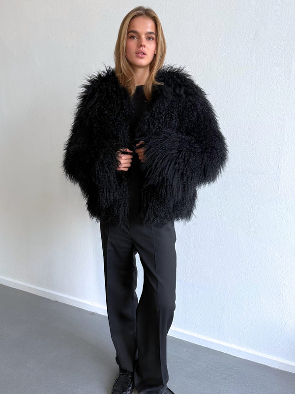 A model showcases a stylish black faux fur jacket by MEOTINE, paired with sleek black trousers against a minimalist backdrop.