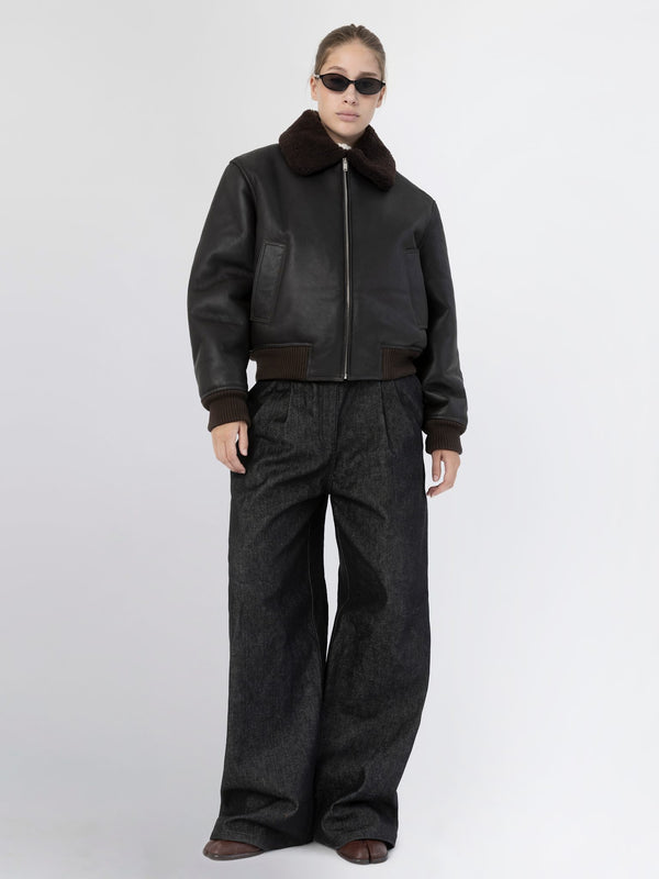 A model showcases a stylish black leather bomber jacket with a brown fur collar, paired with wide-leg dark denim pants. MEOTINE.