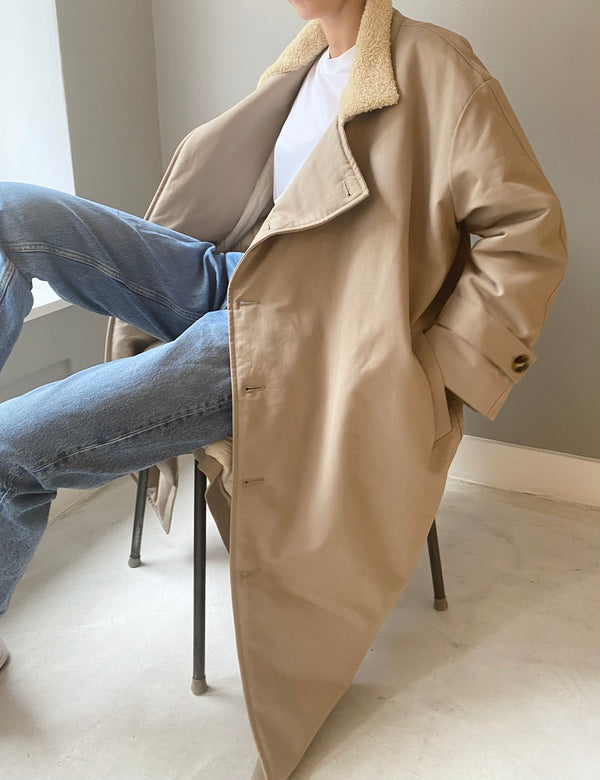 A person sits casually in a chair, wearing a beige MEOTINE coat with a soft collar, paired with light blue jeans.