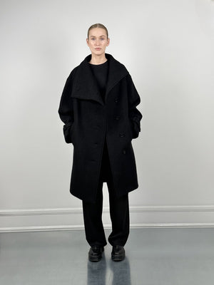 A model showcases a stylish black oversized coat by MEOTINE, paired with a sleek black outfit against a minimalist backdrop.