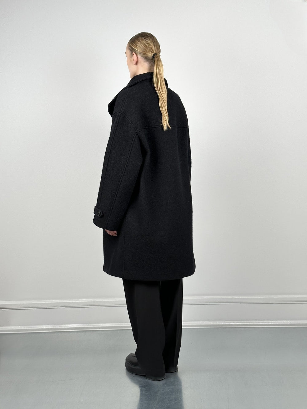 A model showcases a stylish black oversized coat by MEOTINE, paired with a sleek black outfit against a minimalist backdrop.