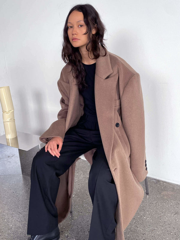 A model sits elegantly in a spacious brown oversized coat by MEOTINE, paired with a black outfit, showcasing a modern, chic style.