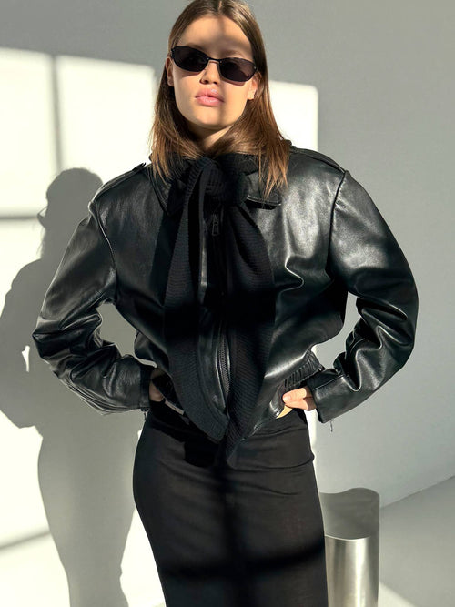 A model wearing a stylish black leather jacket and sunglasses, showcasing a chic look with a sleek black skirt. MEOTINE.