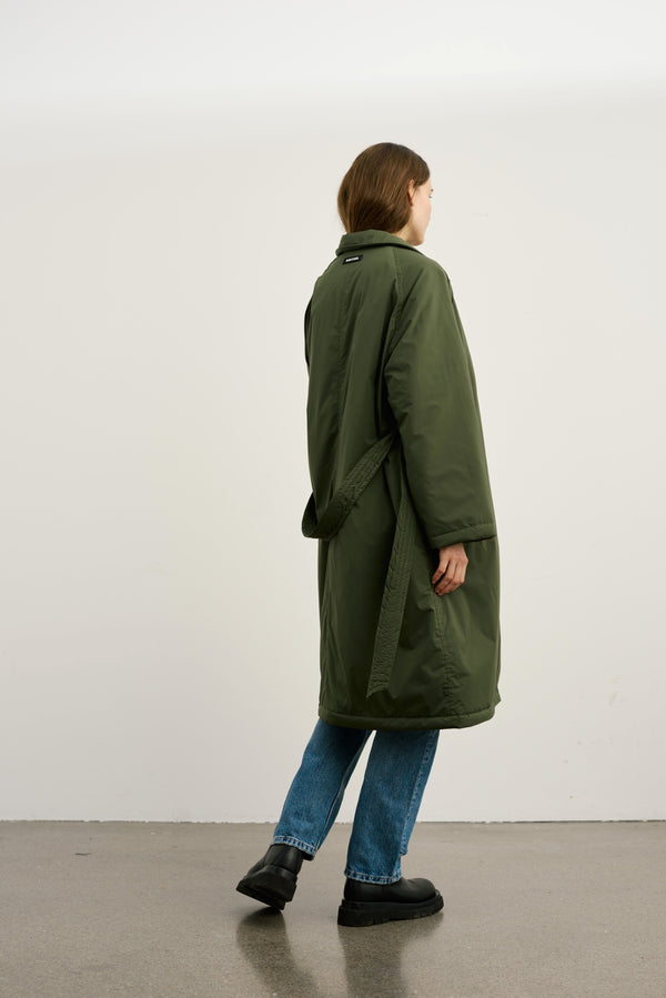 A model wearing a long, olive green MEOTINE coat, paired with blue jeans and black boots, stands against a plain background.