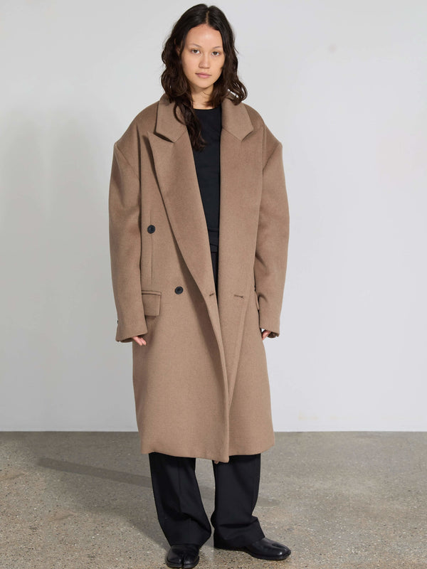 A model showcases a stylish oversized brown coat by MEOTINE, paired with a black top and loose black pants, set against a minimalist backdrop.