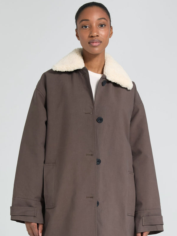 A model wears a brown MEOTINE coat with a plush white collar, showcasing a relaxed fit and button-up design against a neutral background.